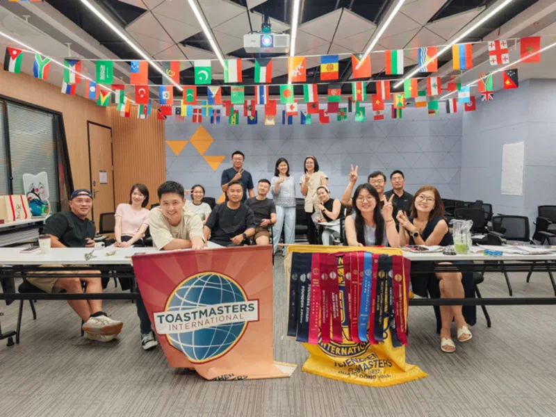 Members and guests of Shenzhen Toastmasters behind the club banner, flags of many countries strung across the ceiling